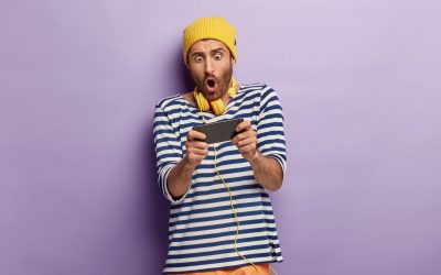 Photo of shocked unshaven guy holds mobile phone horizontally, holds breath, plays games online, being addicted, wears yellow hat and striped jumper, stands in studio against purple background